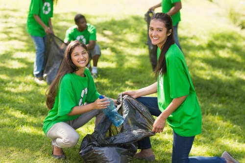 A group of young volunteers outdoors on a bright, sunny day engaged in a community clean-up activity, wearing matching green t-shirts with a recycling symbol. In the foreground, two women are smiling at the camera while holding a large black rubbish bag, which they are either filling or emptying with collected waste. One of the women is kneeling on grass, holding the bag open with both hands, and a clear plastic water bottle is visible nearby. Behind them, three other volunteers, also dressed in green, are actively involved in picking up litter and placing it into similar bags, demonstrating teamwork and environmental effort. The background features a well-maintained grassy area with a few scattered trees providing partial shade, and the overall scene portrays an organized, positive atmosphere of community environmental responsibility. The setting appears to be a park or open space, with natural light casting soft shadows and highlighting the group’s collaborative cleaning activity, supporting local eco-conscious initiatives.