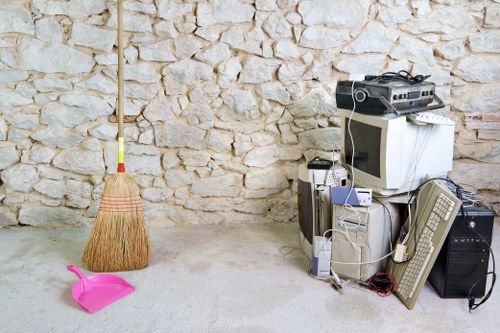 In the foreground, there is a pink plastic dustpan with a curved handle and a matching small broom with natural straw bristles and a yellow handle, positioned on a concrete floor. To the left, a traditional straw broom with a red handle leans against a textured stone wall made of irregularly shaped, light beige stones. On the right side of the image, there is a collection of electronic waste and miscellaneous items stacked together, including an old white CRT computer monitor, a beige tower CPU, a grey keyboard, a black desktop computer case, and various cables and cords. The environment appears to be an indoor or covered storage area, possibly used for rubbish collection or disposal, with neutral lighting highlighting the worn textures of the stone wall and the concrete floor, consistent with the scope of commercial waste removal services in Lee, South East London.
