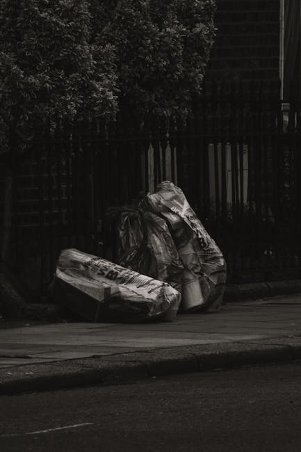 Two large black trash bags filled with waste are placed on a paved sidewalk in front of a metal fence, with a dense cluster of leafy trees behind. The bags appear crumpled and are made of durable plas