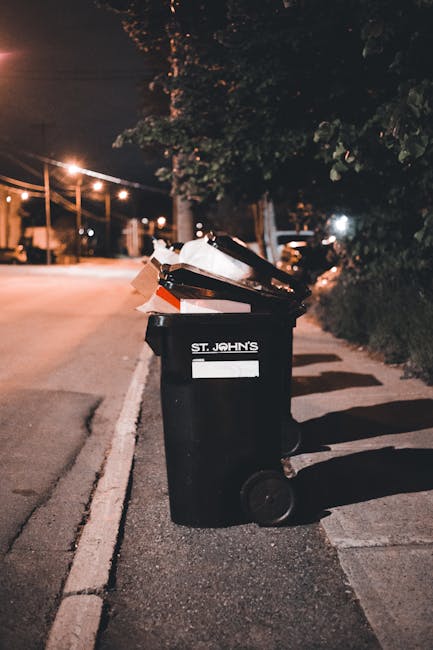 Two large black trash bags filled with waste are placed on a paved sidewalk in front of a metal fence, with a dense cluster of leafy trees behind. The bags appear crumpled and are made of durable plas