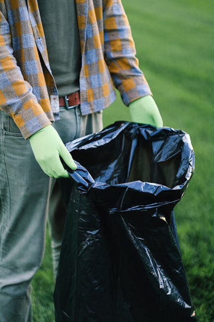 A person wearing a plaid shirt with yellow, grey, and blue tones, paired with green gloves, is holding open the top of a large black rubbish bag made from shiny, durable plastic material. The bag is p
