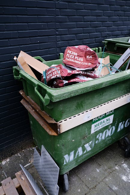 A person wearing a plaid shirt with yellow, grey, and blue tones, paired with green gloves, is holding open the top of a large black rubbish bag made from shiny, durable plastic material. The bag is p